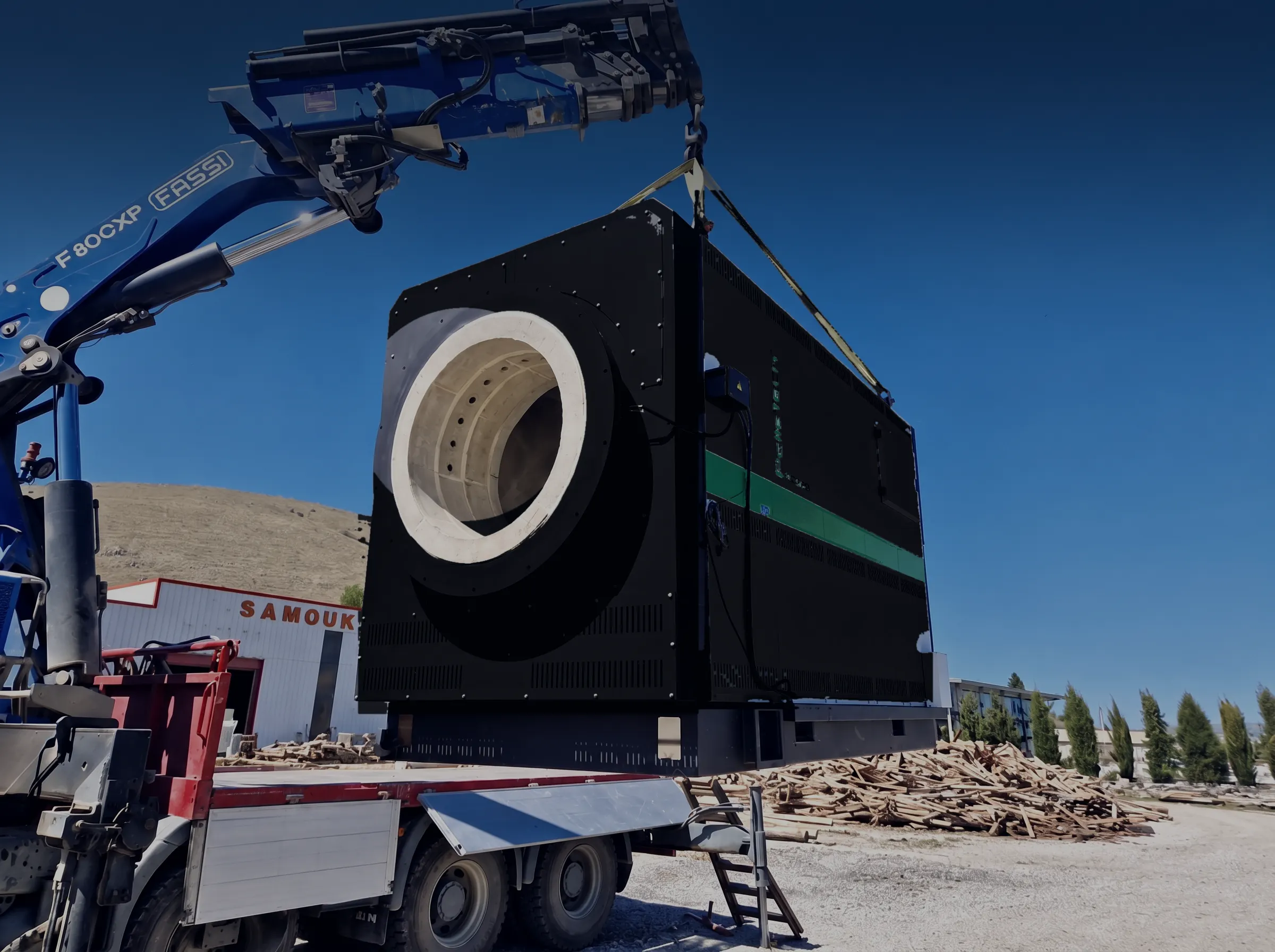A PelleTech BU burner, black with a blue-green stripe, being lifted into place by a truck crane