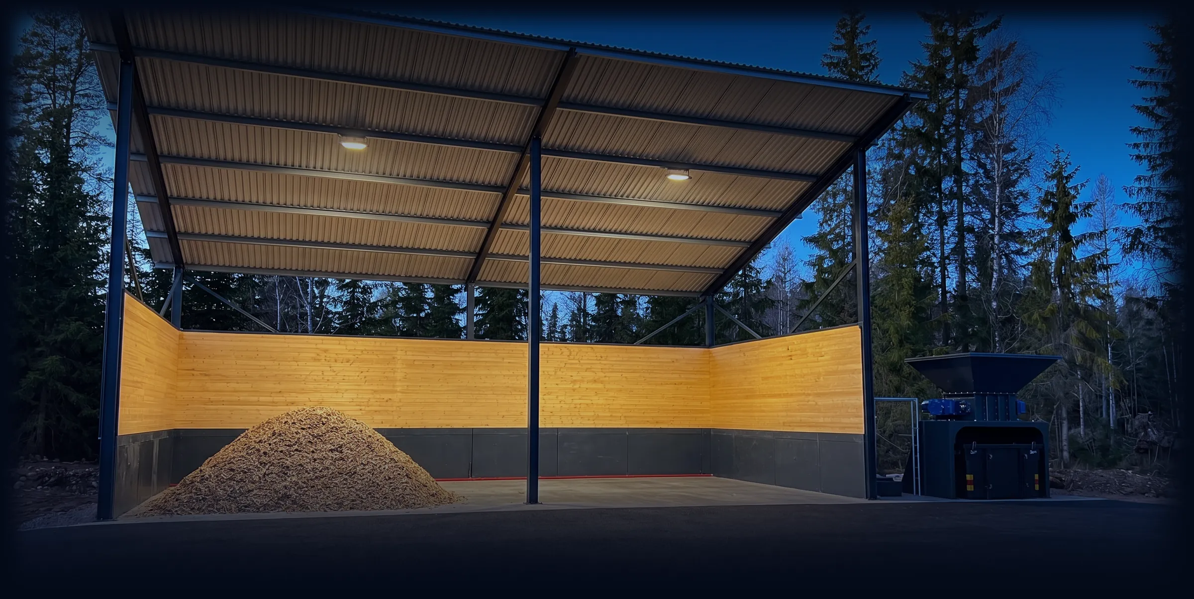Wood chip carport at dusk: a steel frame and sheet-metal roof over a pile of wood chips, with the mobile shredder beside it