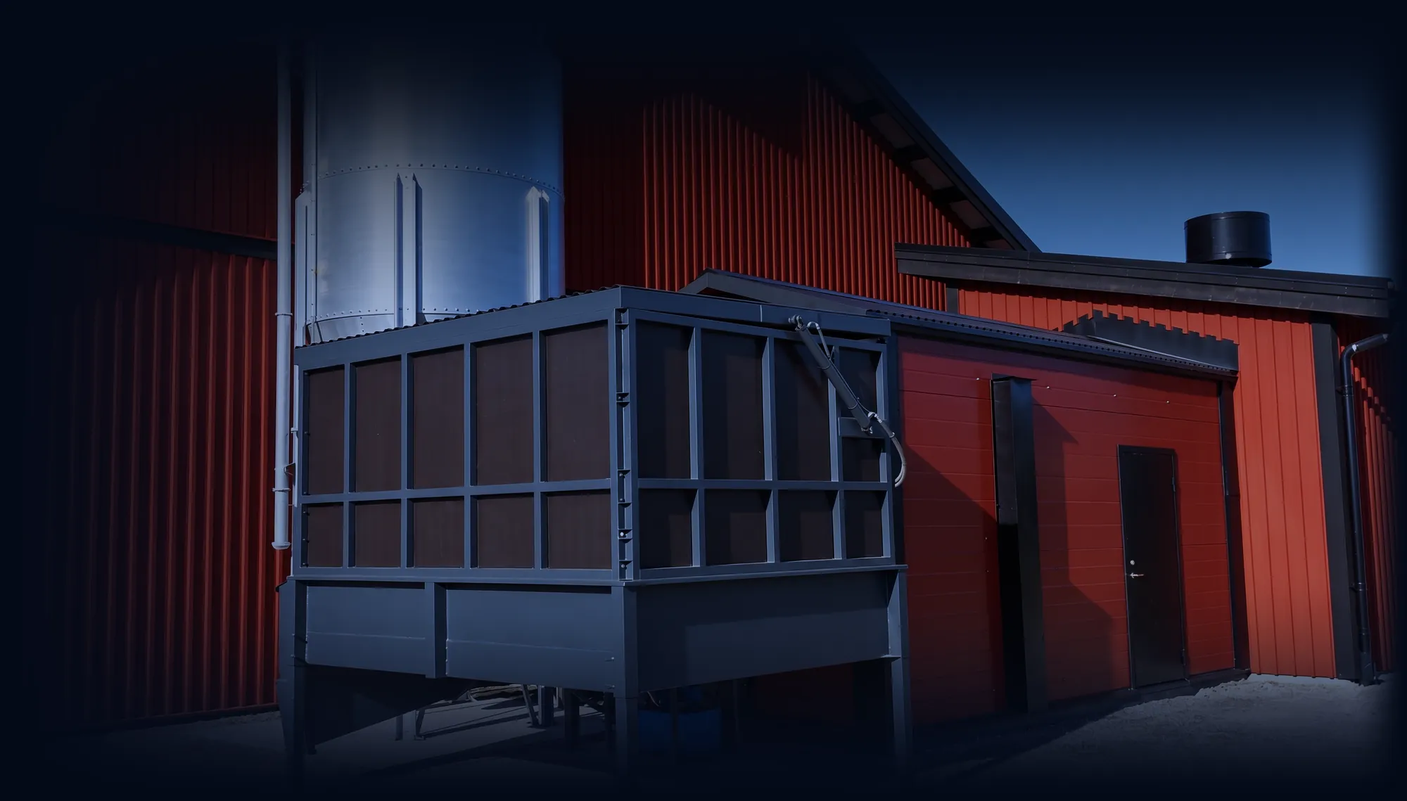 Wood chip fuel bunker on steel legs beside a red industrial building with a silo, at dusk
