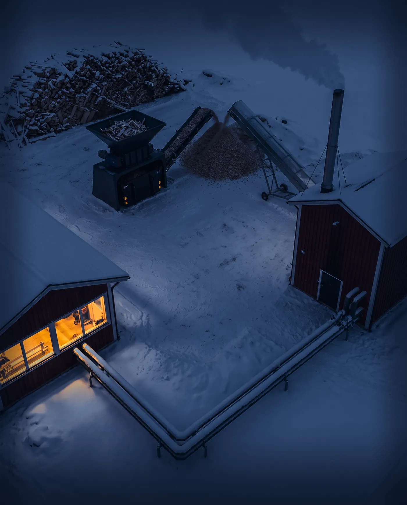 A farm in snow, seen from above at dusk: a pile of wood offcuts, the Shredder, a pile of wood chips by a conveyor belt, the boiler house with its chimney and the pipes to a lit workshop