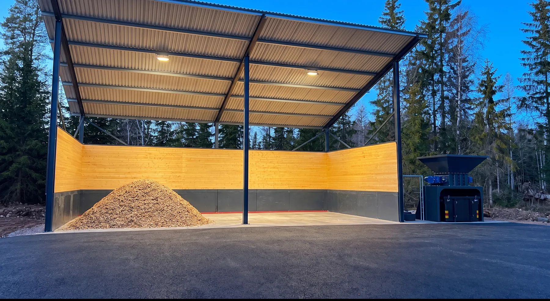 The Shredder next to a covered wood chip store, a pile of chips in the store and forest behind it, one evening