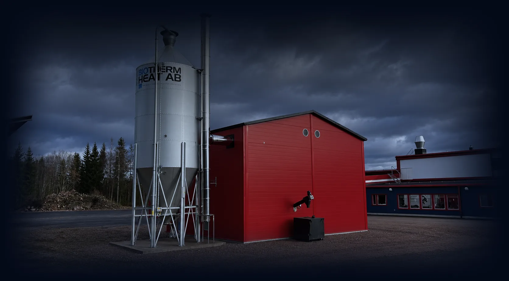 Galvanised pellet silo with the Biotherm Heat logo beside a red boiler house, in evening light