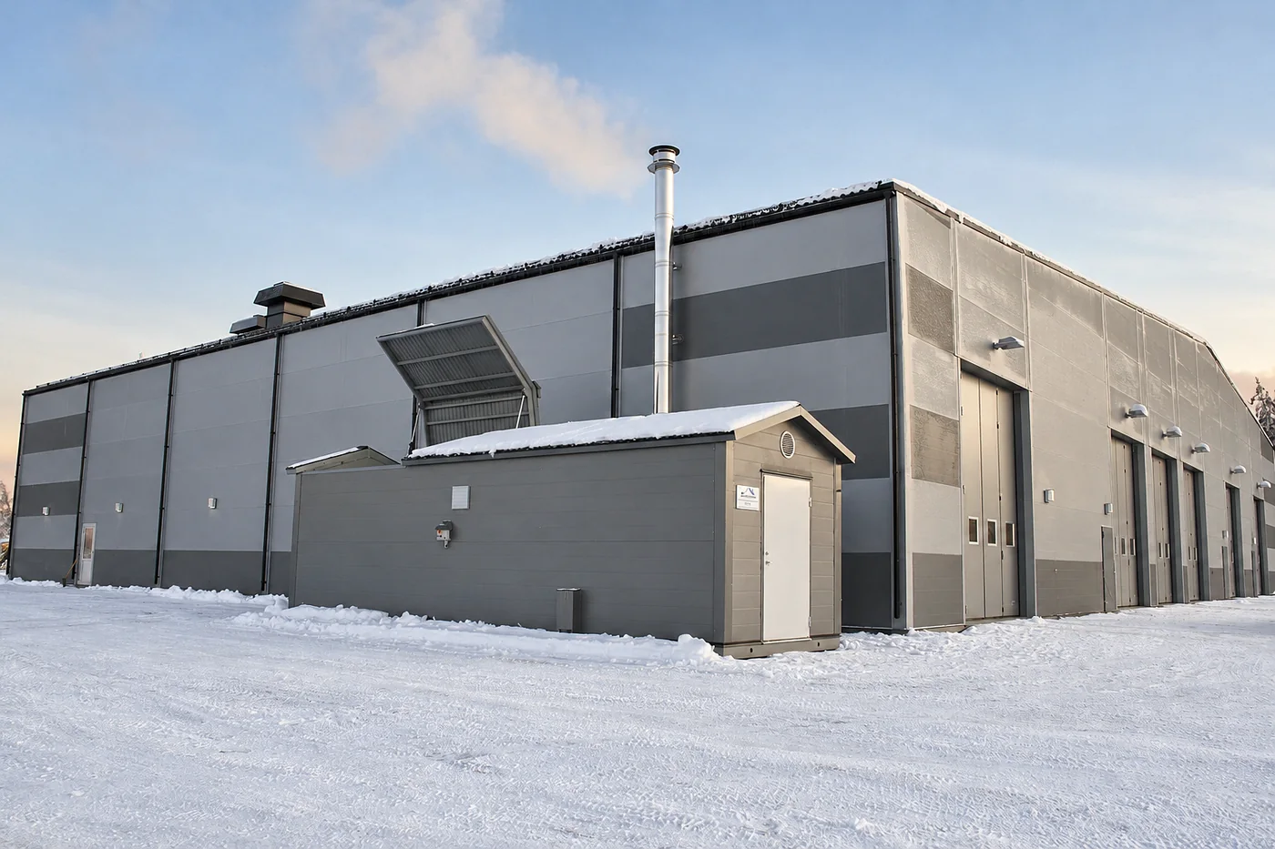 Grey boiler house with the roof hatch raised and a chimney, next to a larger hall in winter snow